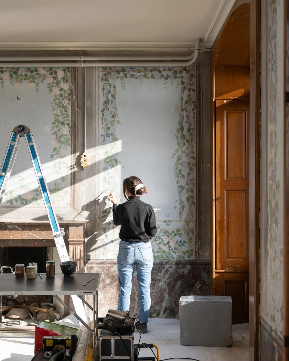 A conservator-restorer treating a trompe-l’œil wall painting at the Domaine de La Doges, in a sunlit historic interior.